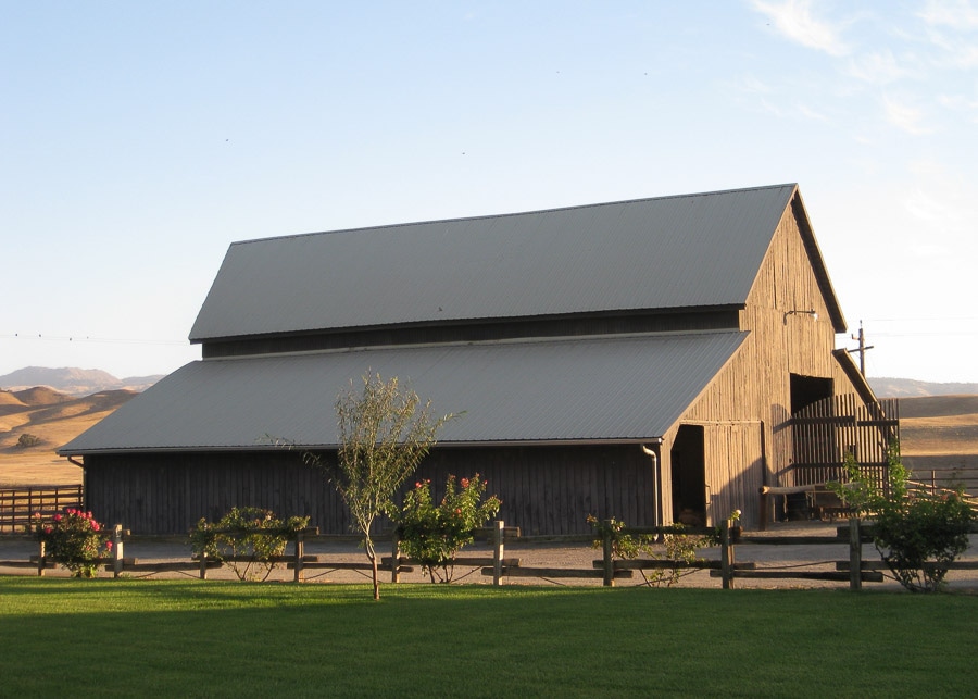 A large brown barn with a wood fence, roses, and green grass surrounding it.