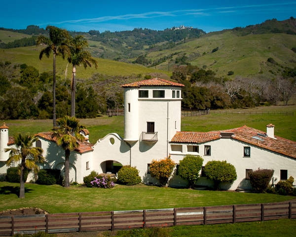 A spanish style building sits among green hills on the San Simeon Ranch. Hearst Castle is in the distant background.