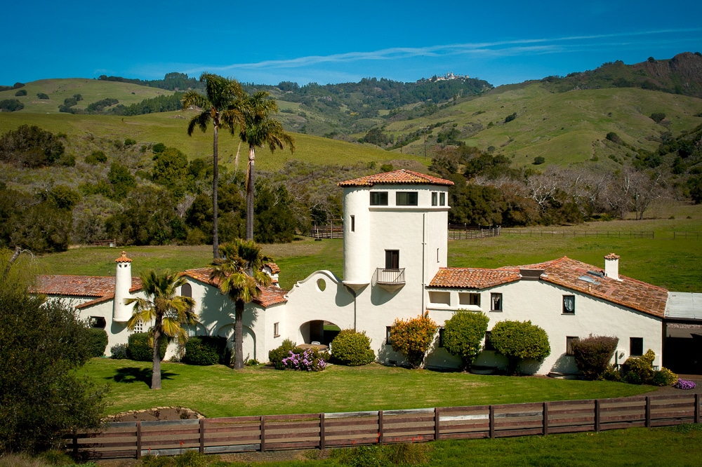 A spanish style building sits among green hills on the San Simeon Ranch. Hearst Castle is in the distant background.