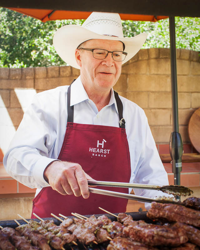 Steve Hearst wearing a cowboy hat and red apron with the Hearst Ranch Beef logo on it. He is holding tongs and grilling skewered meat on a grill.