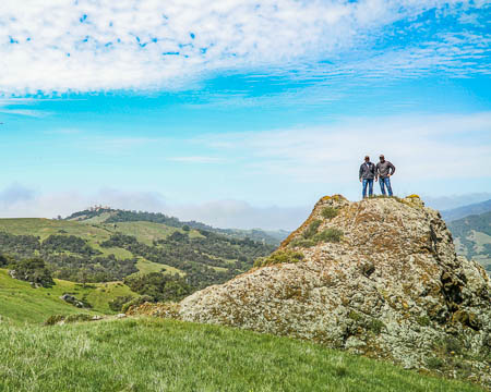 Two men standing on a rocky hill, with Hearst Castle in the background.