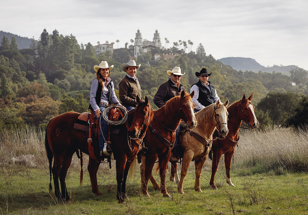 Four Hearst Ranch team members on horseback, standing side by side with the Hearst Castle in the background.