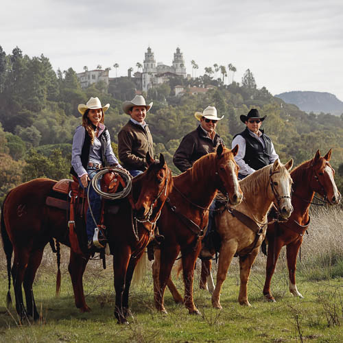 Four Hearst Ranch team members on horseback, standing side by side with the Hearst Castle in the background.