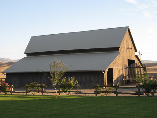A large brown barn with a wood fence, roses, and green grass surrounding it.