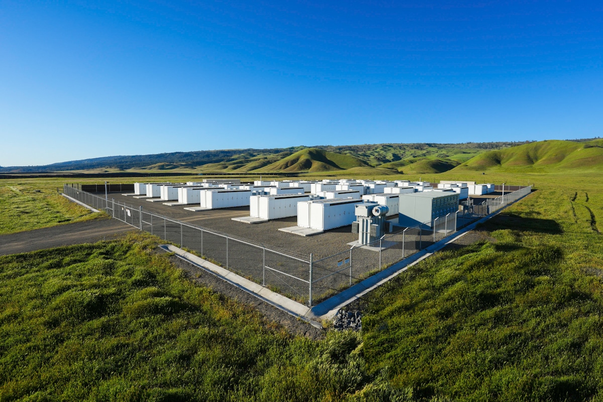 An aerial shot of a Tesla Megapack battery station, surrounded by green grass and green hills.