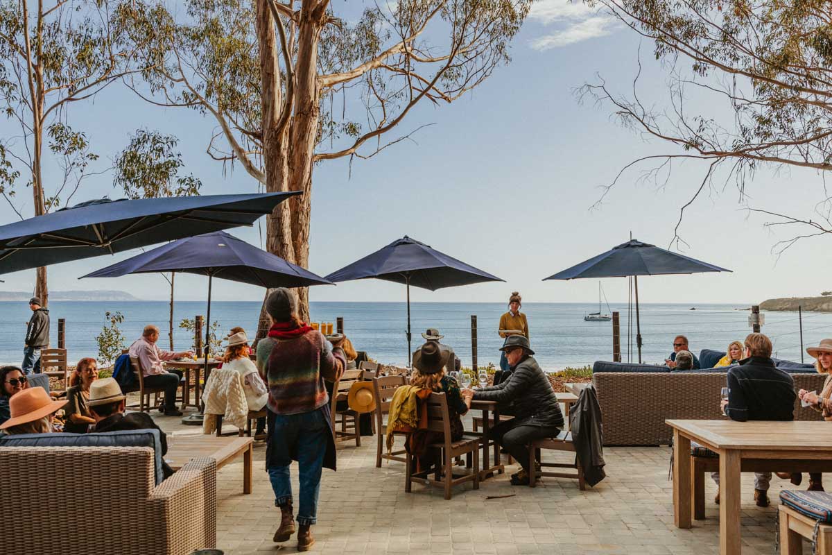 Hearst Ranch Winery outdoor tasting room with wicker and wood chairs and tables, blue umbrellas, and a view of the ocean.