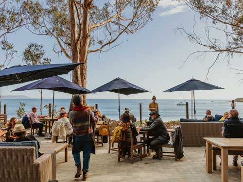 Hearst Ranch Winery outdoor tasting room with wicker and wood chairs and tables, blue umbrellas, and a view of the ocean.